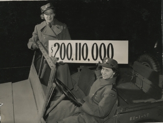 Deux femmes soldats dans une jeep de l’armée, tenant une pancarte indiquant $200,110, 000.