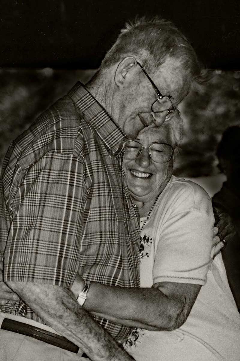 Un homme tient sa femme dans une étreinte aimante et elle sourit largement.