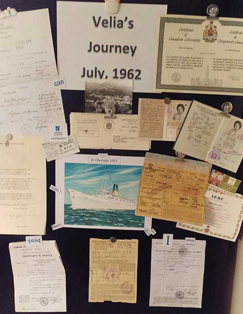 Plusieurs anciens documents, cartes postales et photos sont épinglés sur un tableau d’affichage avec le titre Velia’s Journey 1962.