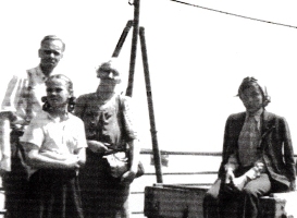 Quatre personnes sur le pont à bord d’un navire, des poulies à l’arrière-plan.