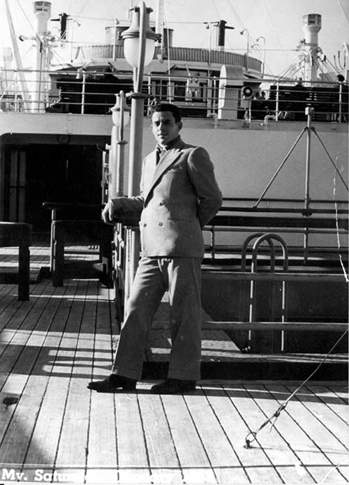 Un jeune homme en costume se penche sur la rambarde d’un navire.