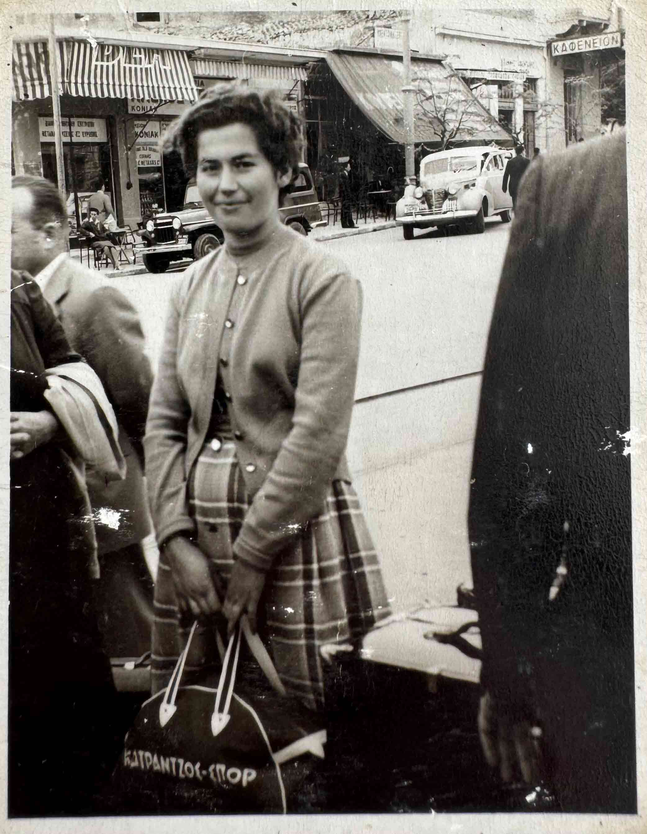 Photographie en noir et blanc d’une femme grecque souriant et tenant un bagage.