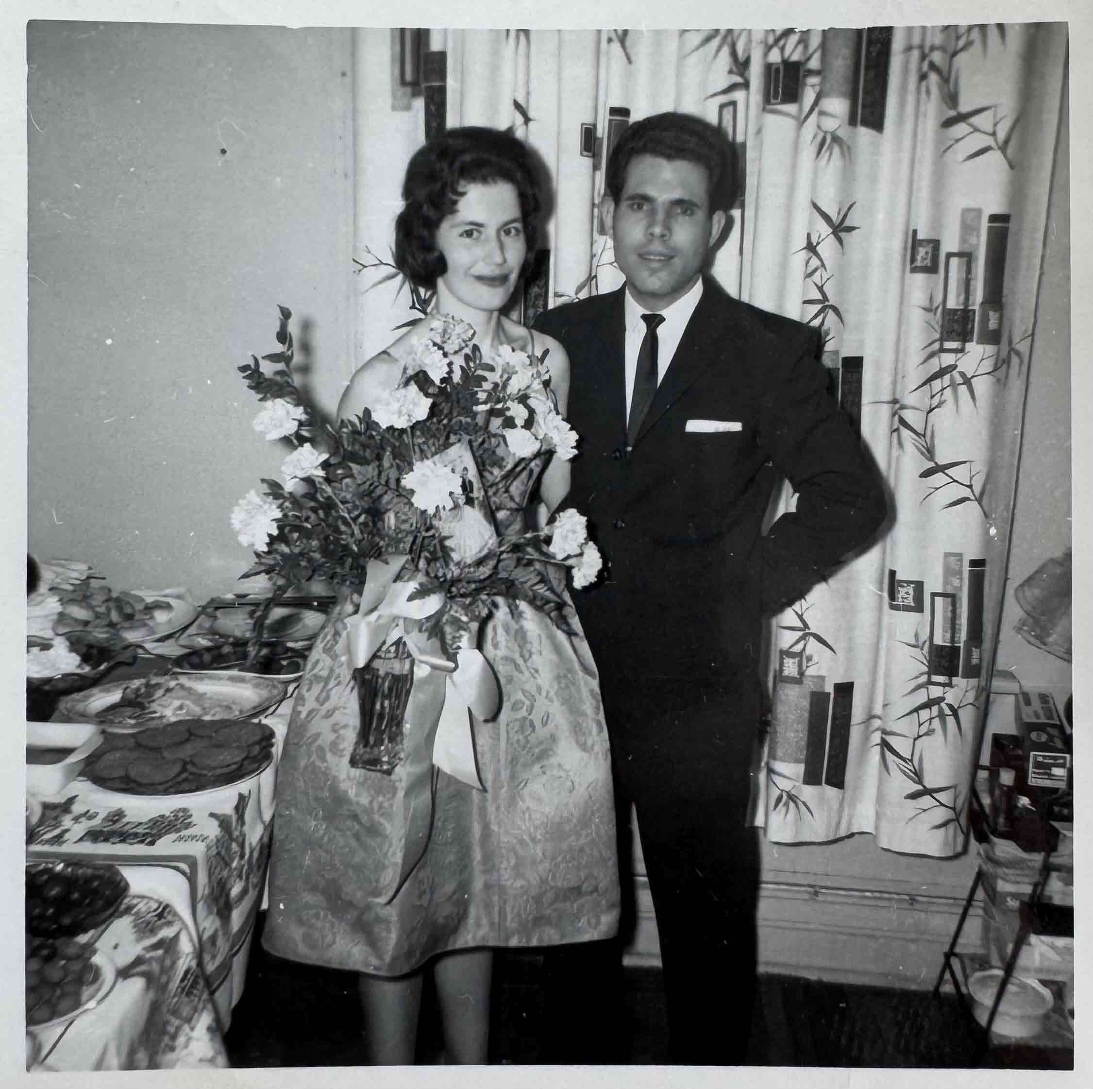 Photographie en noir et blanc d’un couple grec en tenue de cérémonie, avec des fleurs.