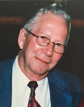 Portrait d’un homme blanc âgé et souriant portant des lunettes, un complet bleu et une cravate à motifs rouges.