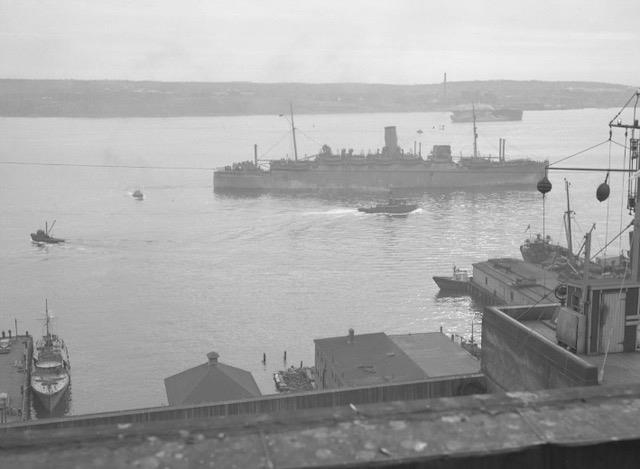 Photo en noir et blanc d’un grand navire dans un port, avec des bateaux plus petits autour de lui.