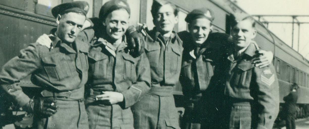 Photo en noir et blanc de cinq hommes blancs souriants en uniforme, bras dessus, bras dessous, avec un train en arrière-plan.