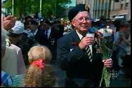 Un homme blanc, âgé et souriant, en uniforme, portant un bouquet de fleurs, marchant dans une foule de personnes.