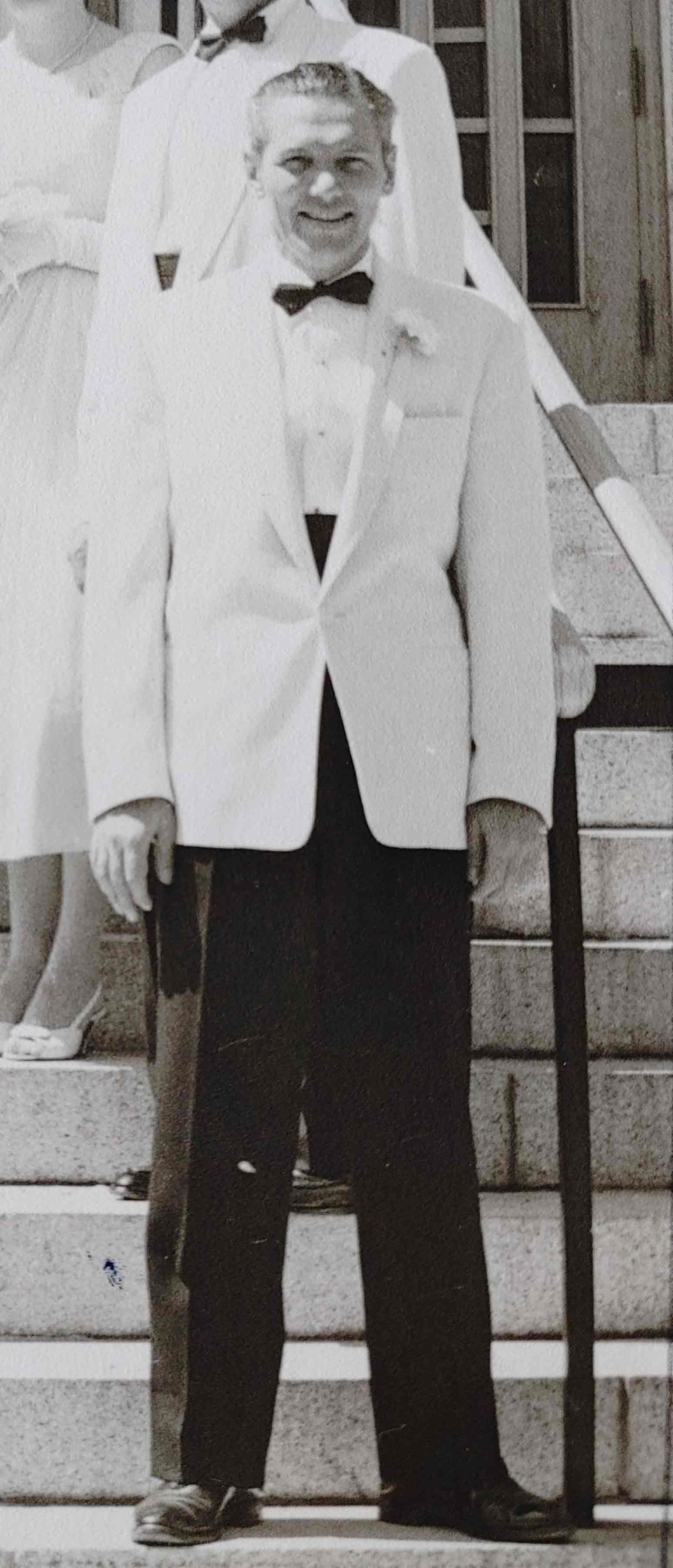 Photo en noir et blanc d’un homme blanc vêtu d’un pantalon noir et d’un veston blanc, debout sur des marches, avec d’autres personnes derrière lui.