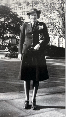 Photo en noir et blanc d’une femme blanche souriante en uniforme, debout à l’extérieur d’un grand bâtiment.