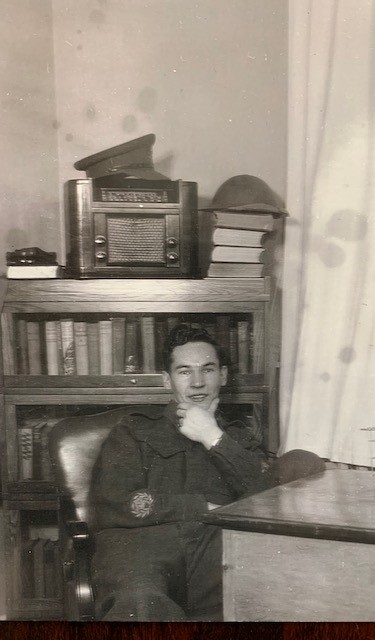 Photo en noir et blanc d’un homme blanc souriant, le menton dans la main, assis à un bureau avec une bibliothèque derrière lui.