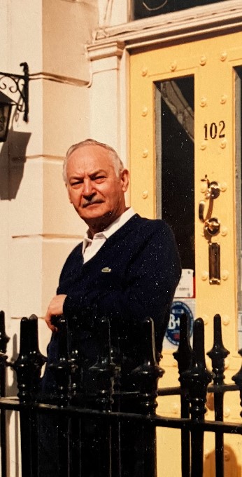 Un homme blanc âgé devant une porte d’entrée, un bras appuyé sur une clôture noire.